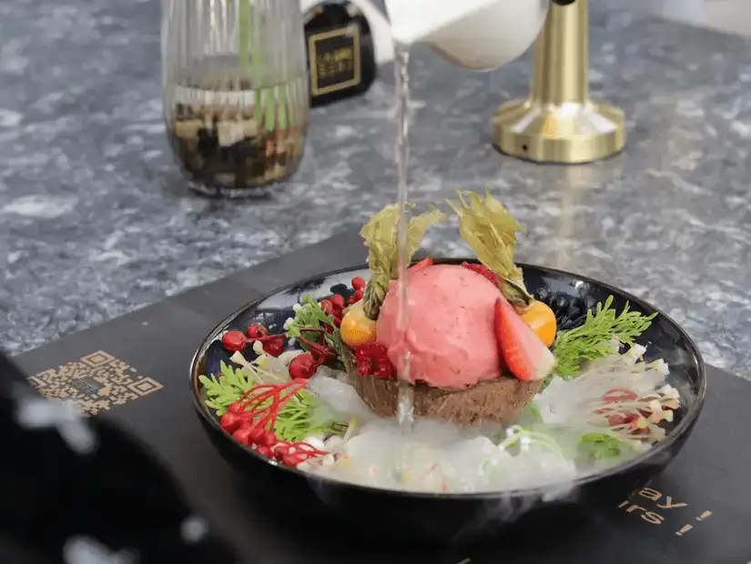 An artistically presented dessert featuring a scoop of pink ice cream with a liquid pour, adorned with fresh berries and leaves.