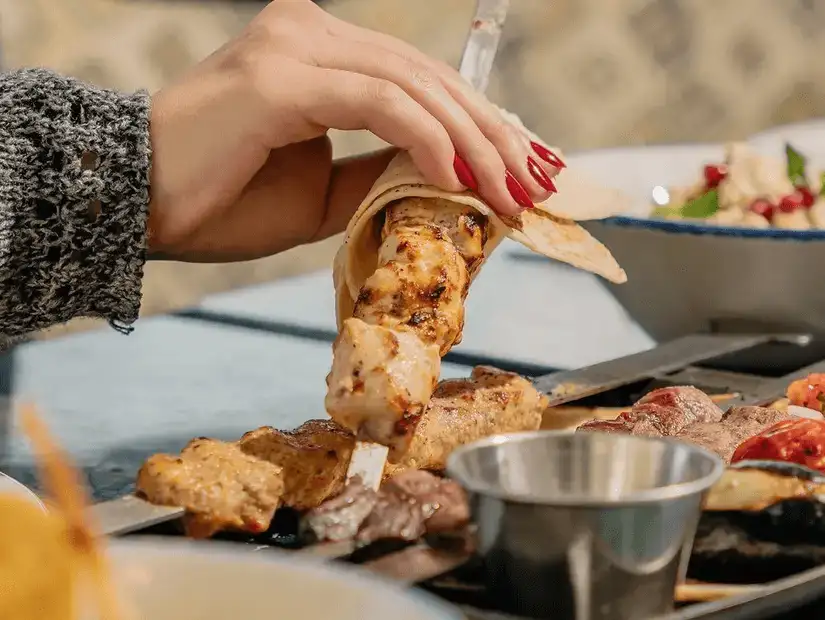 A hand holding a piece of grilled chicken kebab wrapped in flatbread, with an assortment of meats and vegetables in the background.