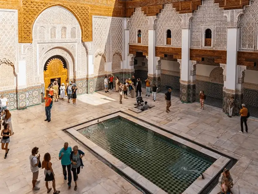 Tourists explore a Moroccan courtyard with intricate architecture and a reflecting pool.