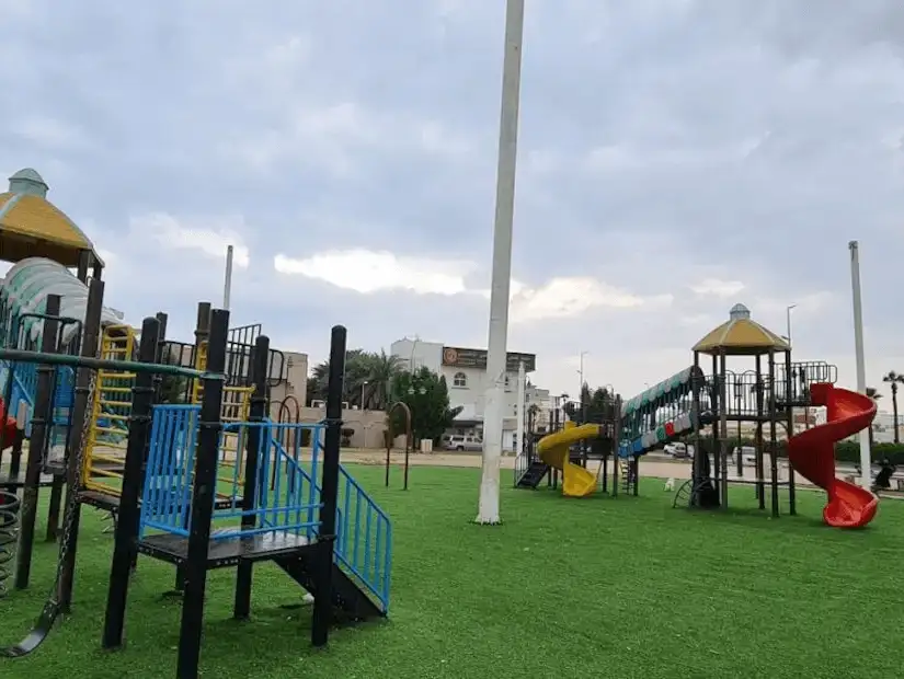 Colorful children's play area with slides and climbing frames on a soft grassy surface in Saleh Kamel Park.