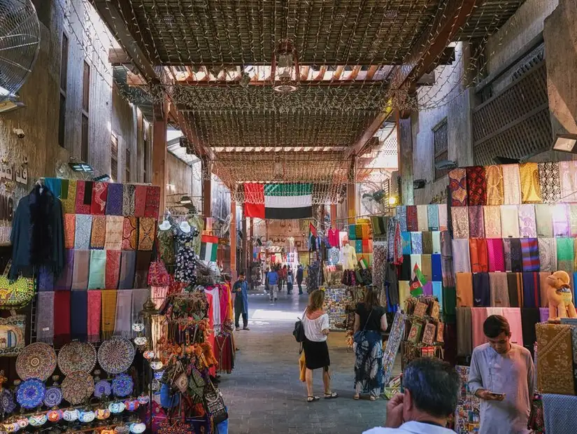 A colorful alley under a protective canopy, filled with rich fabrics and lively trade.