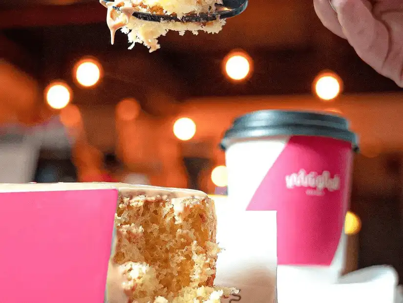 A slice of cake being lifted, paired with a bright pink take-out coffee cup, in a cozy cafe setting with warm lighting.