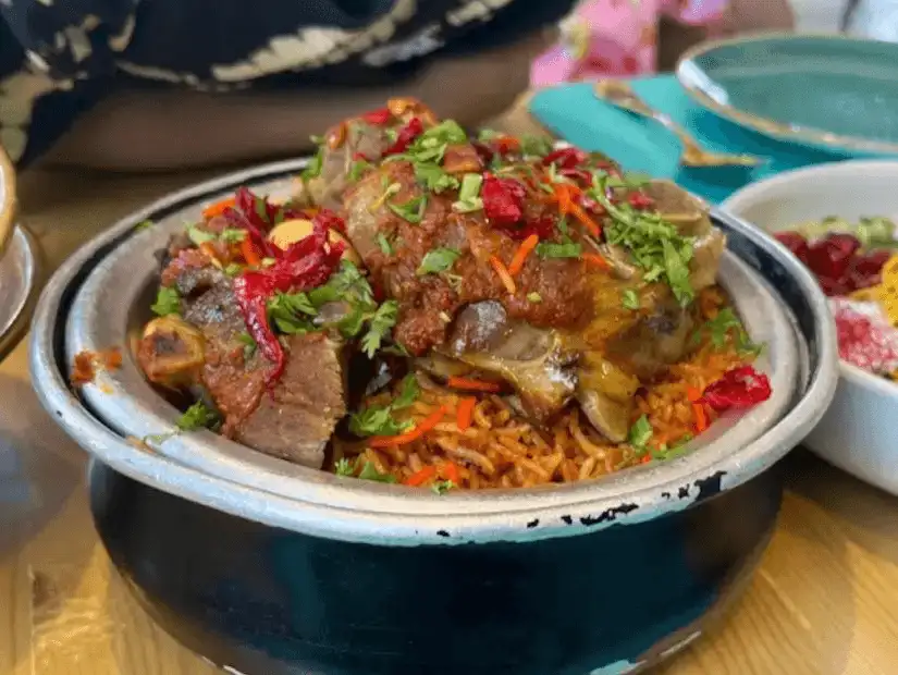 A steaming pot of spiced rice with lamb, garnished with fresh herbs, red peppers, and almonds, ready for a communal meal.