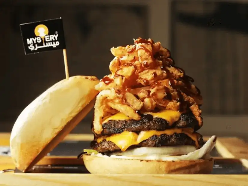 A towering cheeseburger stacked with crispy fried onions and drizzled with sauce, branded with a 'Mystery' flag.