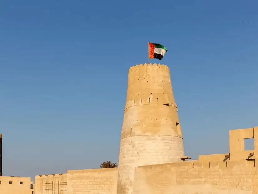 A cylindrical, sand-hued tower topped with the UAE flag rises over the historic coastal settlement’s fort walls.