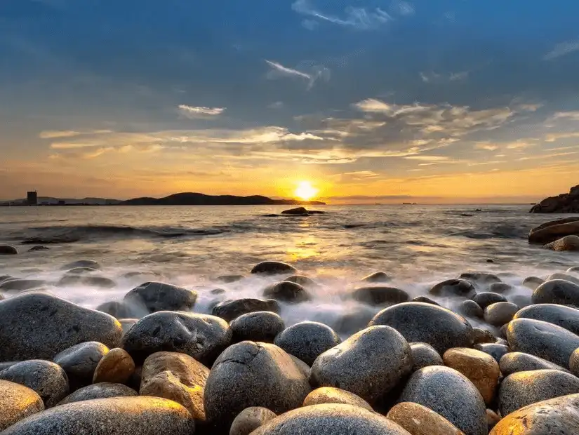 A picturesque rocky shoreline with waves gently lapping against the stones during a golden sunset.