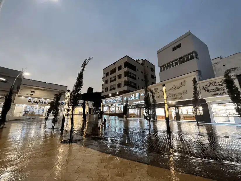 A wet street in Al-Baha with shops and buildings illuminated at dusk.