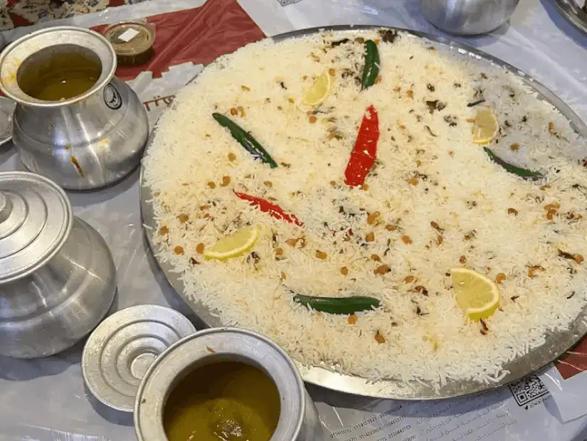 A traditional rice dish served with green chili, red pepper, and slices of lemon, garnished with fried nuts, and accompanied by sauces.