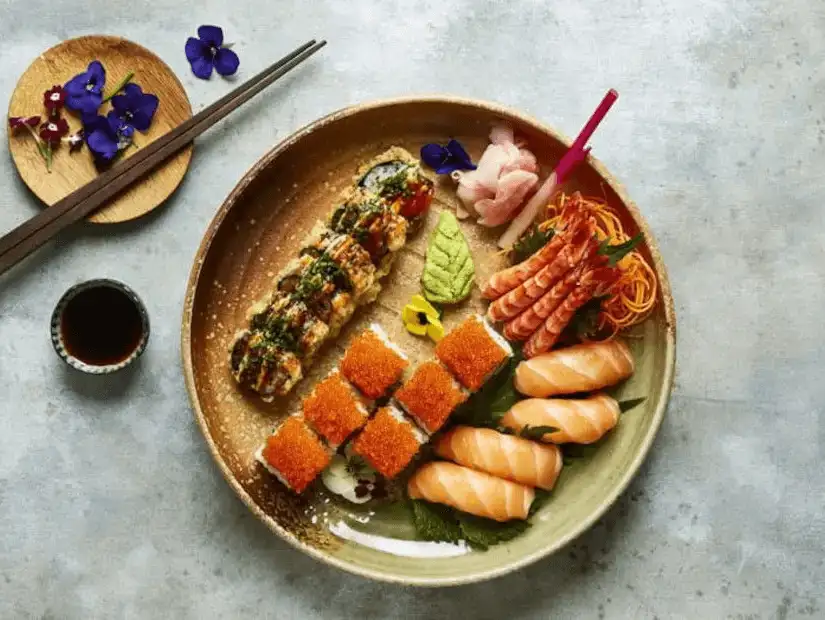 A colorful platter of assorted sushi, sashimi, and rolls, artfully presented with edible flowers and dipping sauce.