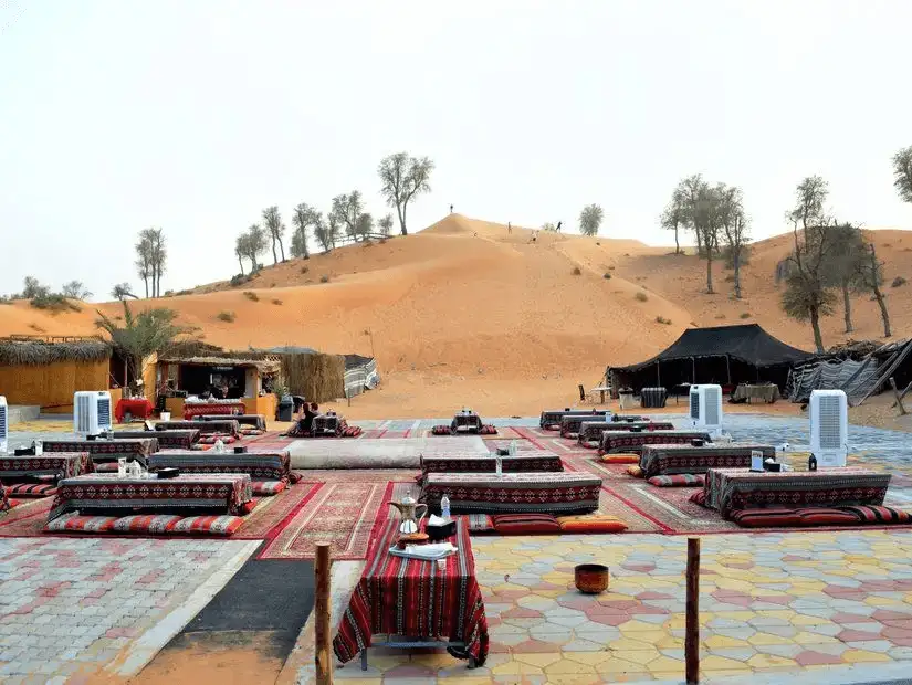 Colorful floor cushions and patterned rugs form open-air seating beside sweeping dunes and traditional tents at a desert camp.