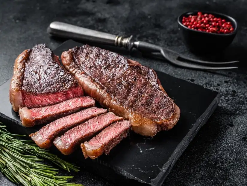 Richly seared steak slices on a black stone slab, complemented by fresh rosemary and vibrant red peppercorns.