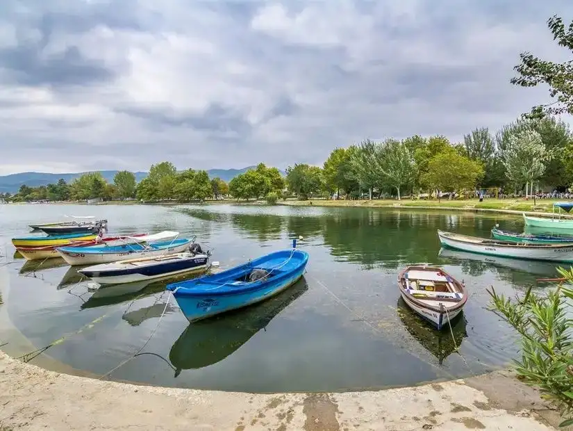 A tranquil lake with colorful boats moored along the shore, surrounded by green trees and a cloudy sky.