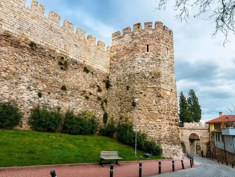 A historic stone wall and tower from an ancient castle in Bursa, standing against a cloudy sky with green gardens around.