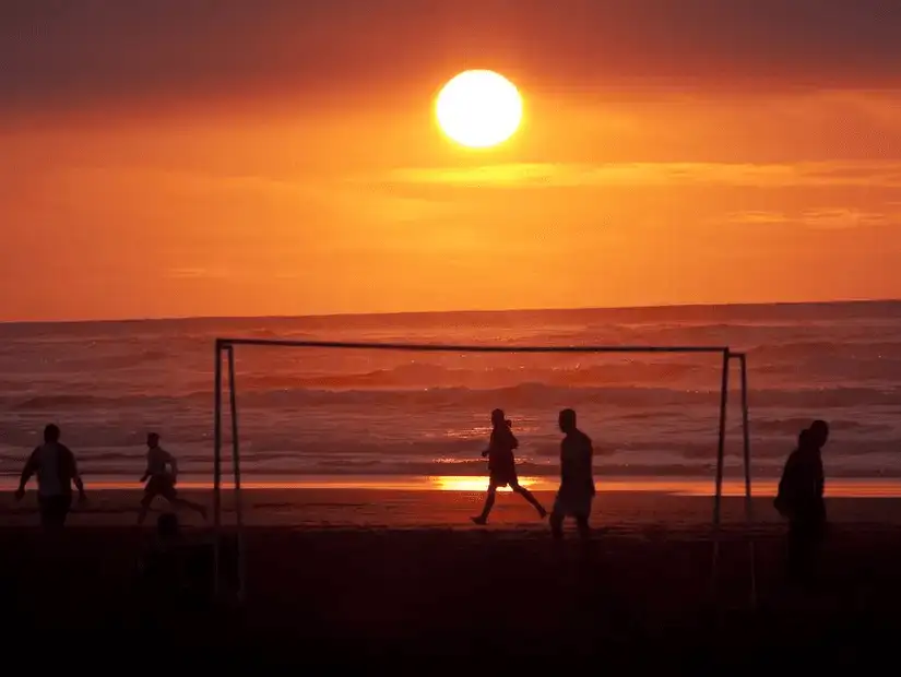 Silhouetted figures playing soccer on the beach at sunset.