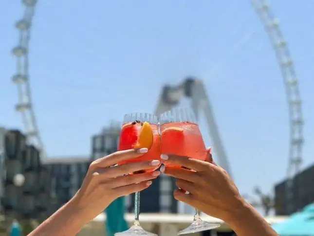 Two people toasting with fruity cocktails in front of a Ferris wheel at the beach.