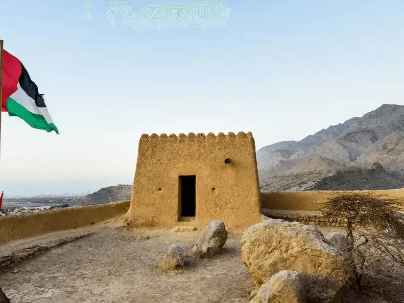 An ochre watchtower with crenellated roof stands on a rocky ridge beside a fluttering UAE flag, backed by rugged mountains.