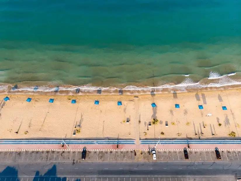 Aerial view of turquoise waves lapping a wide sandy shore dotted with neat rows of blue sunshades beside an empty seaside road.