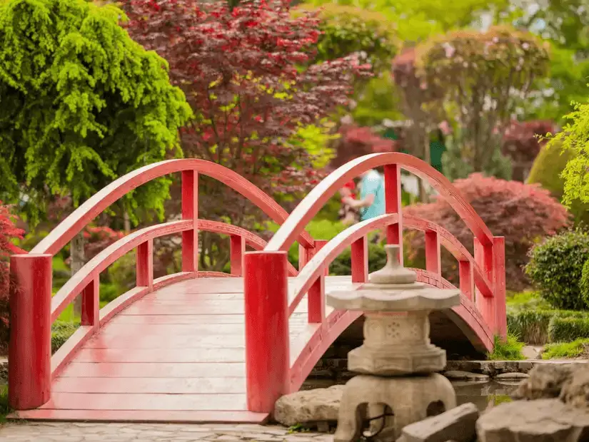 A red bridge in a tranquil Japanese-style garden, surrounded by vibrant greenery.