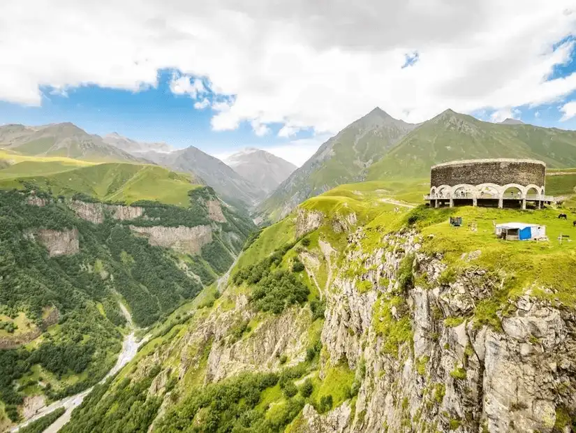 A scenic mountainous landscape with a historic round structure perched on a cliff.