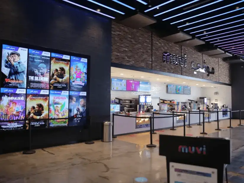 The lobby of a cinema with large digital screens displaying movie posters and a snack counter, creating a lively cinematic experience.