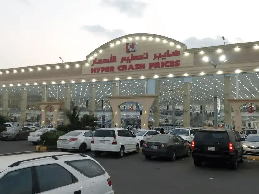 A large and brightly lit supermarket with shoppers and parked cars in front.