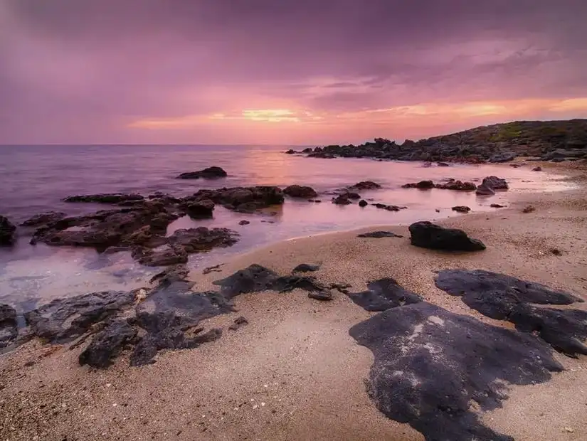 A serene coastal sunset at Jabal Hassan, where soft purple hues meet the gentle sea against a rocky beach.