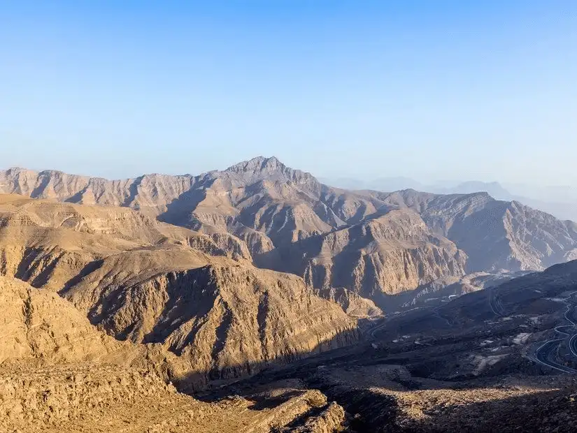 Rugged, sun-baked ridges cascade toward hazy horizons from the UAE’s highest mountain peak under a clear sky.