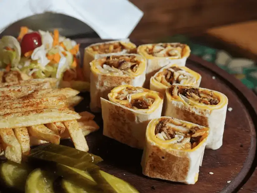 A platter featuring rolled wraps with tender meat, served with fries, pickles, and salad.