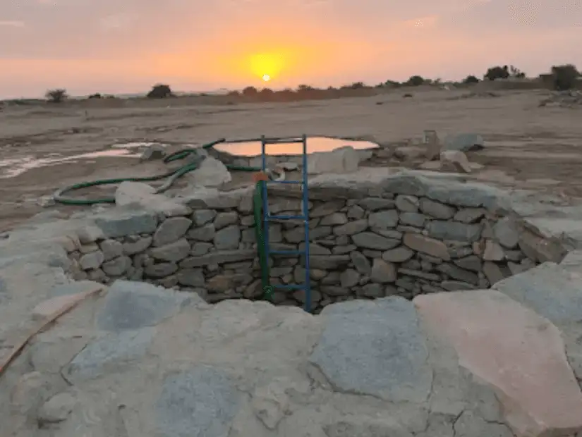 A stone well with a ladder at sunset, set in a barren landscape.