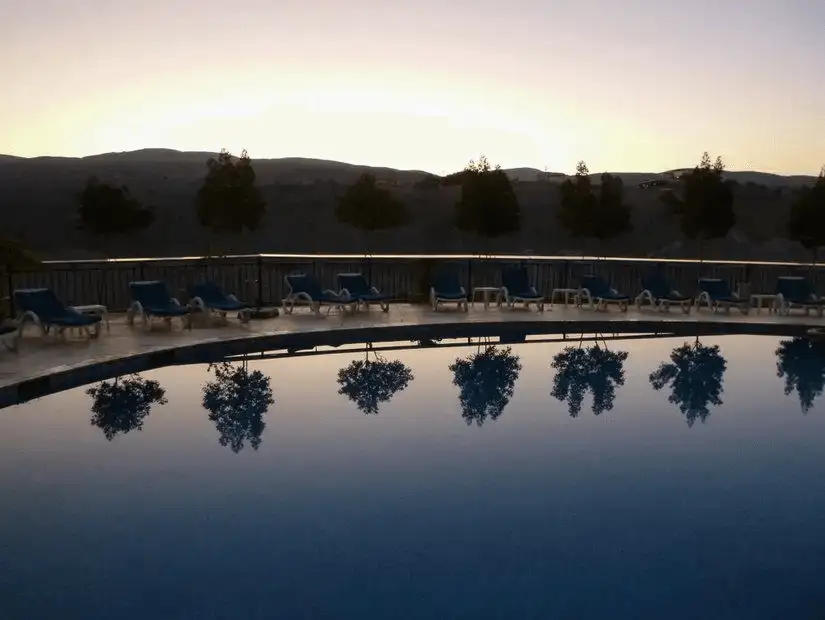 Twilight silhouettes trees around a tranquil, mirror-smooth thermal pool reflecting the fading glow of surrounding hills.