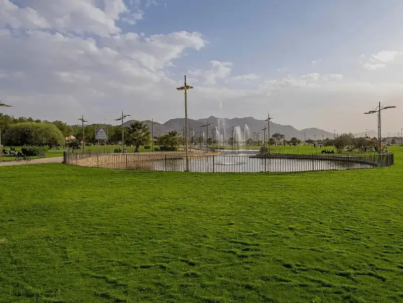 A serene view of King Fahd Park, featuring lush green lawns and a central fountain under a bright sky.