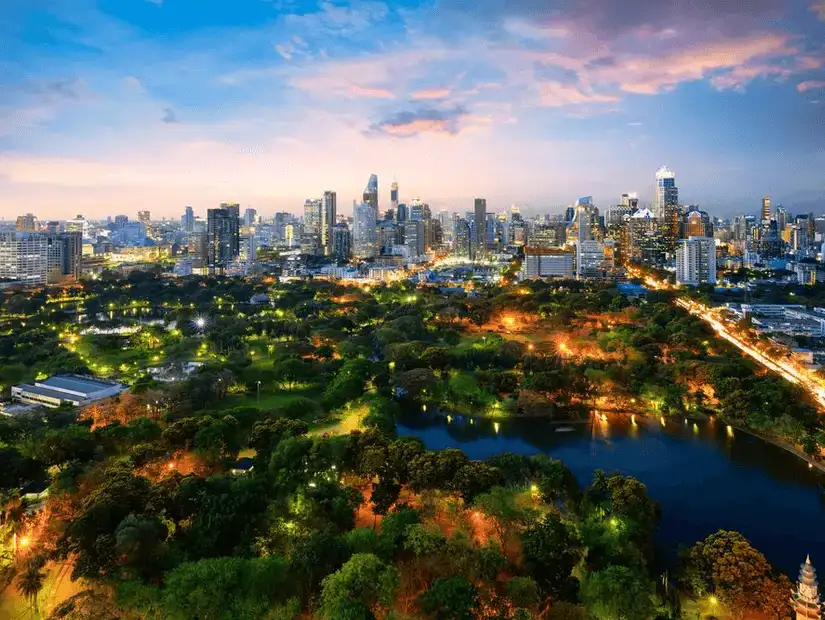 A vibrant skyline seen from the sprawling greenery of Lumpini Park at dusk.