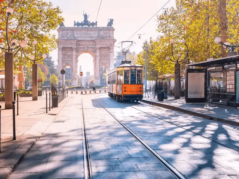 A historic orange tram passing through the quiet streets of Milan with the iconic Arch of Peace in the background.