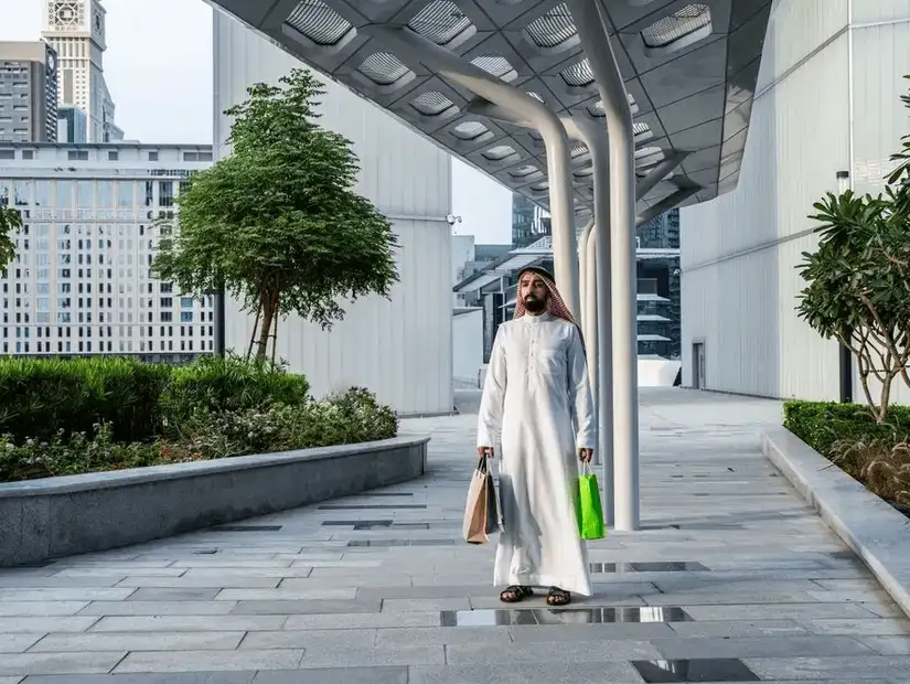 A man in traditional attire carrying shopping bags, walking along a modern cityscape with green spaces and architectural columns.