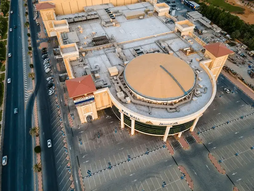 Drone shot of a vast, circular-roofed mall with columned façade and sprawling empty parking lots along a city highway.