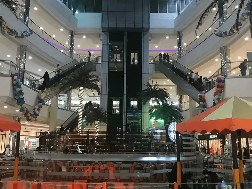 The interior of a modern shopping mall with escalators and palm trees.