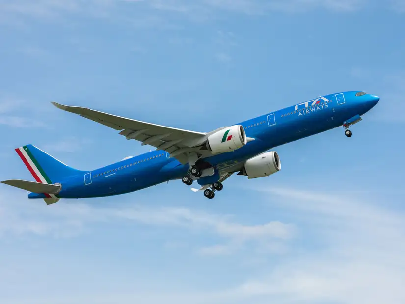 A blue aircraft from ITA Airways taking off with a dramatic sky in the background.