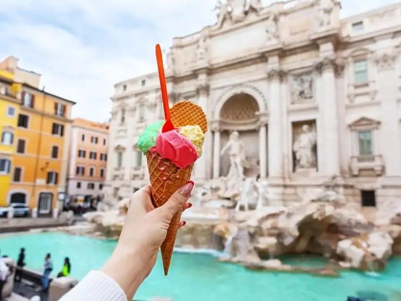 A hand holding a colorful gelato cone in front of the Trevi Fountain in Rome.