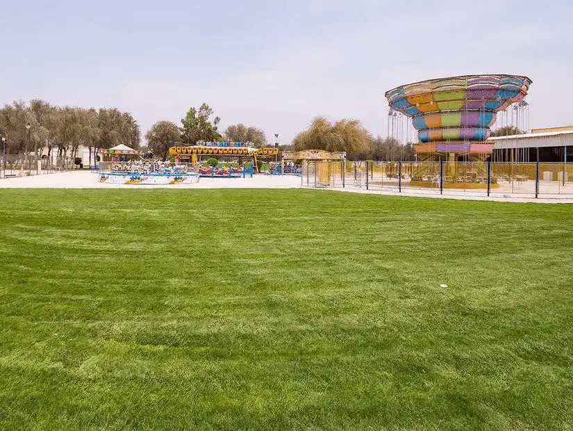 A broad carpet of emerald lawn fronts colorful amusement rides and a towering swing carousel in Saqr Park.