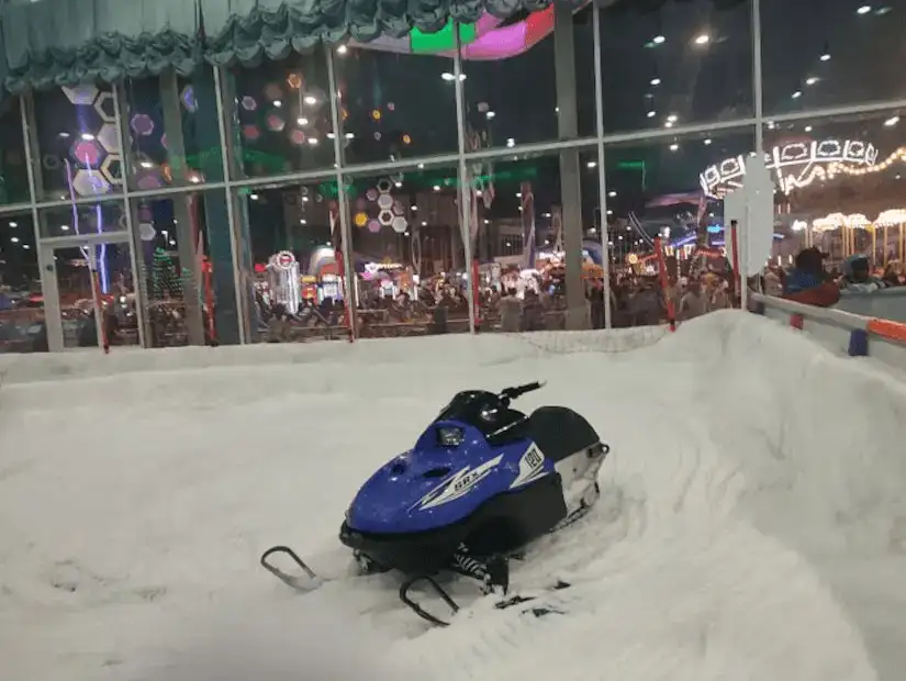 A blue snowmobile sits in an indoor snow park, with lights and rides visible in the background.