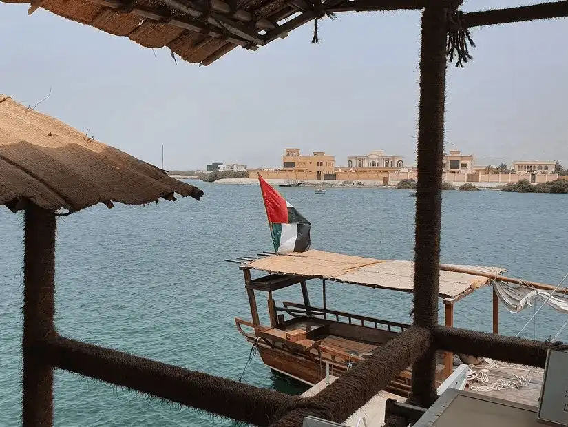 From a thatched pearl-farm hut, a traditional dhow bearing the UAE flag floats on calm coastal waters.