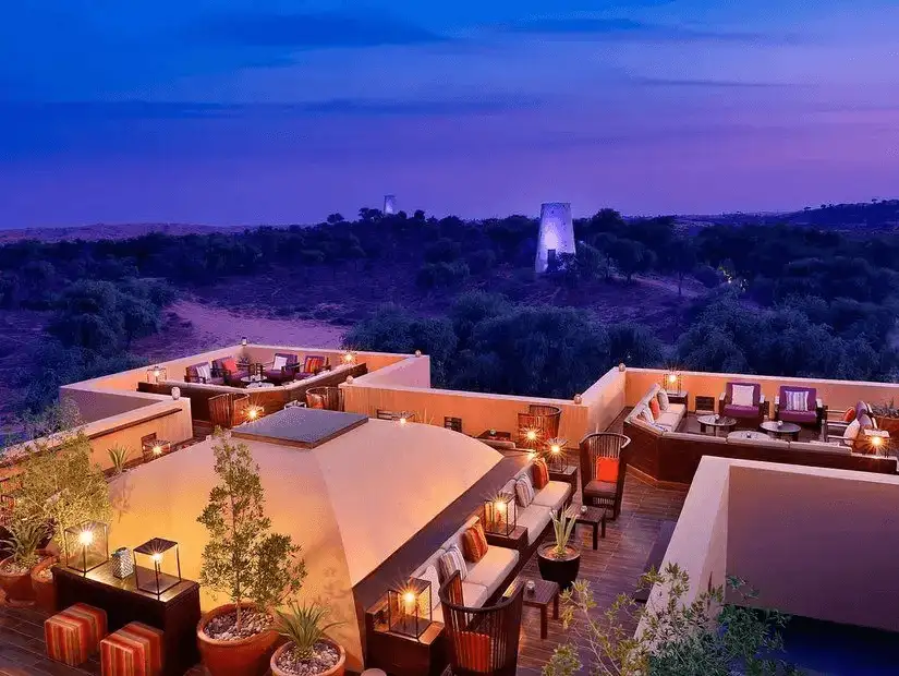 Candle-lit rooftop terraces of a desert resort glow warmly against twilight dunes and distant watchtowers.